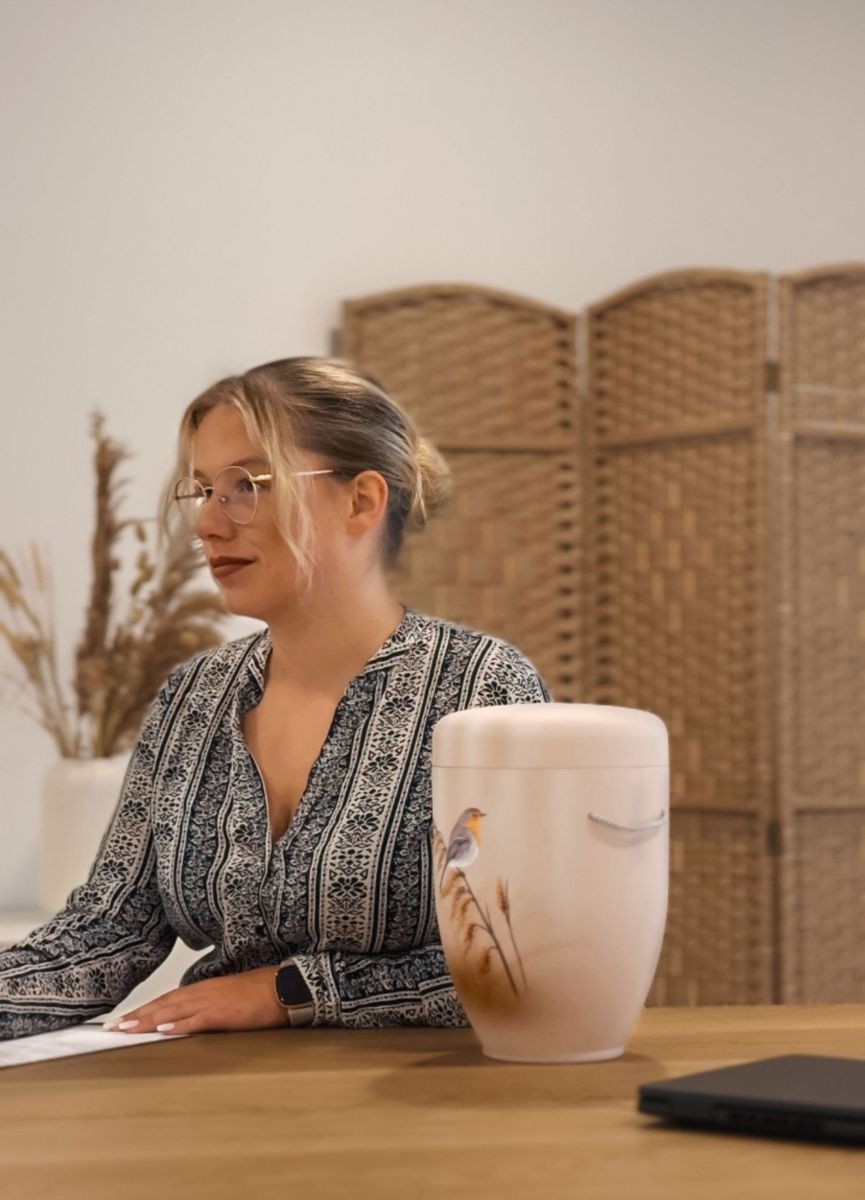 Person in gemustertem Hemd sitzt an einem Holztisch mit einer dekorativen Urne und einem gewebten Raumteiler im Hintergrund.