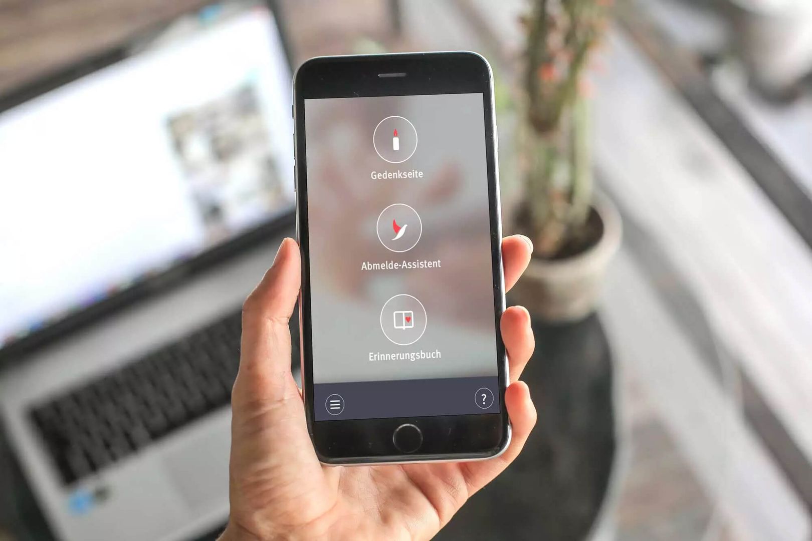 Eine Hand hält ein Smartphone mit einer deutschen App-Oberfläche, im Hintergrund ein verschwommener Laptop und eine Pflanze.